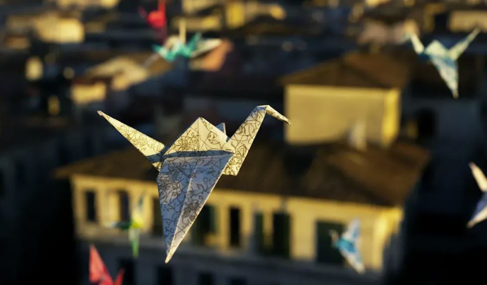Una foto en primer plano de un pájaro de origami volando sobre un paisaje urbano, en una bandada con otros de diferentes colores y patrones, proyectando sombras intrincadas sobre los edificios de abajo.