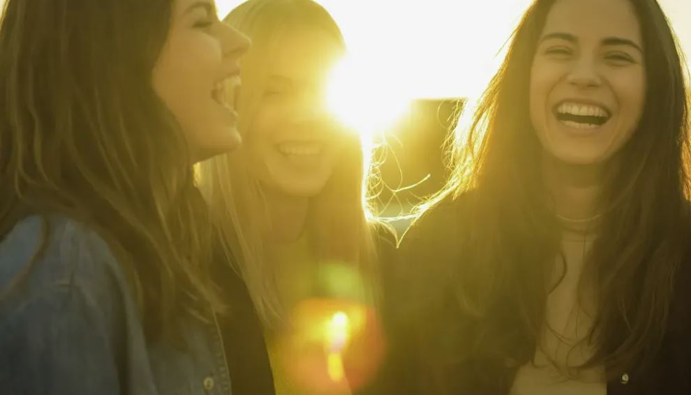 Tres mujeres están juntas riendo, con una mujer ligeramente desenfocada en primer plano. El sol se está poniendo detrás de las mujeres, creando un destello de lente y un brillo cálido que resalta su cabello y crea un efecto bokeh en el fondo. El estilo de fotografía es espontáneo y captura un momento genuino de conexión y felicidad entre amigas. La cálida luz de la hora dorada aporta una sensación nostálgica e íntima a la imagen.